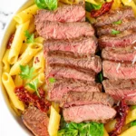 Close-up of a bowl of steak pasta, featuring perfectly cooked penne pasta, sliced medium-rare steak, sun-dried tomatoes, fresh spinach, and grated Parmesan cheese, garnished with fresh parsley and red pepper flakes.