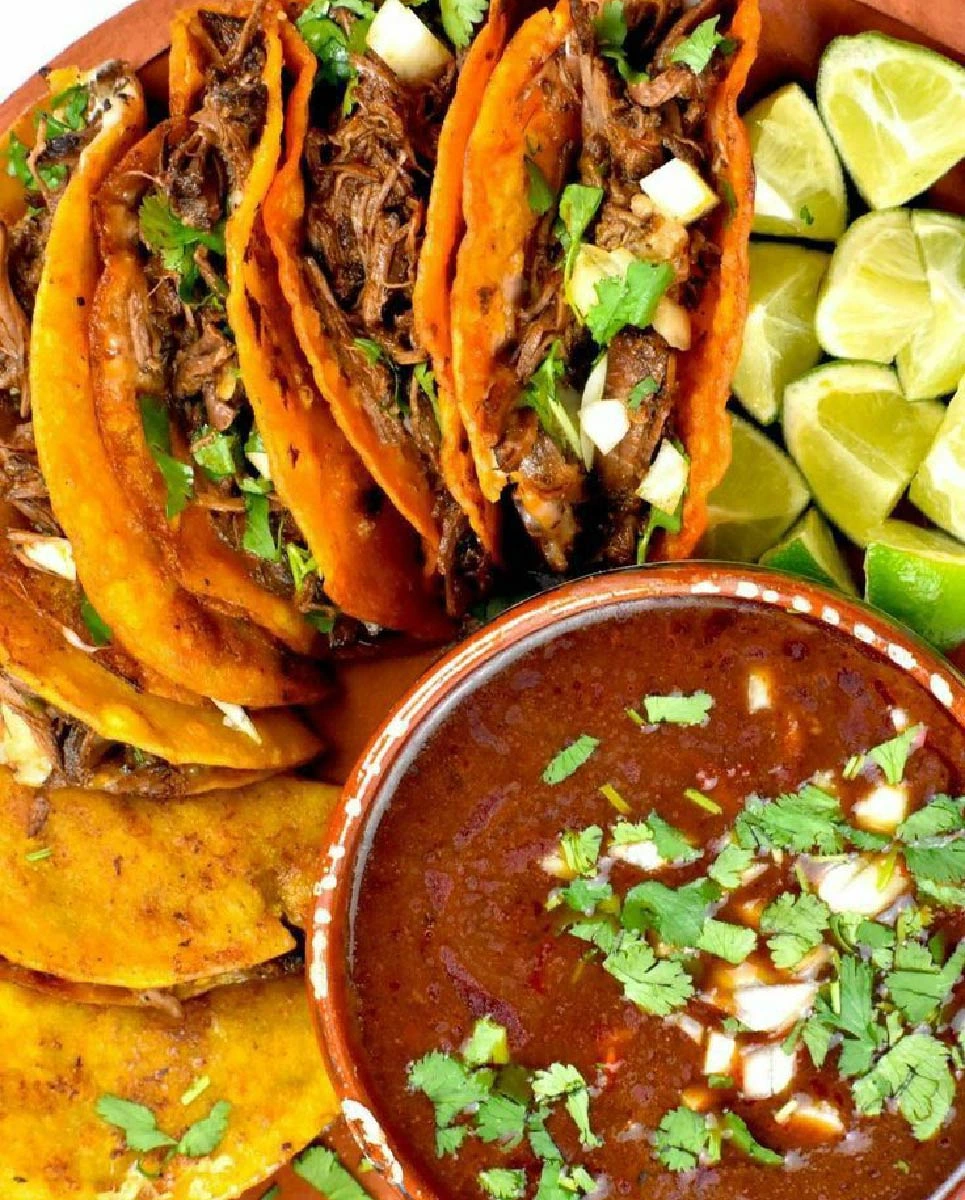 A platter of birria tacos filled with shredded meat, topped with cilantro and onions, served with lime wedges and a bowl of rich consommé garnished with fresh herbs.