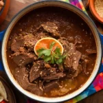 Beef Birria with tender beef in a rich, spicy broth, served with warm tortillas, cilantro, onions, and lime for the perfect bite
