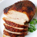 Close-up of a perfectly roasted boneless chicken breast with a crispy, seasoned golden-brown skin, partially sliced to reveal moist white meat, garnished with fresh parsley on a white plate.