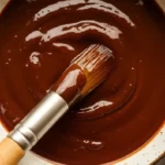 A rustic bowl filled with smooth, glossy BBQ sauce, with a stainless steel and wooden-handled brush resting inside.