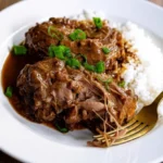 Close-up of tender, braised turkey necks smothered in a rich, thick, brown gravy with visible onions, bell peppers, and fresh green onions in a large cooking pot.