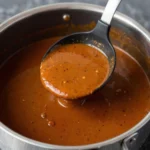 Close-up of rich, brown homemade gravy being ladled from a gleaming stainless steel saucepan into a serving dish, showing its smooth texture and steam.